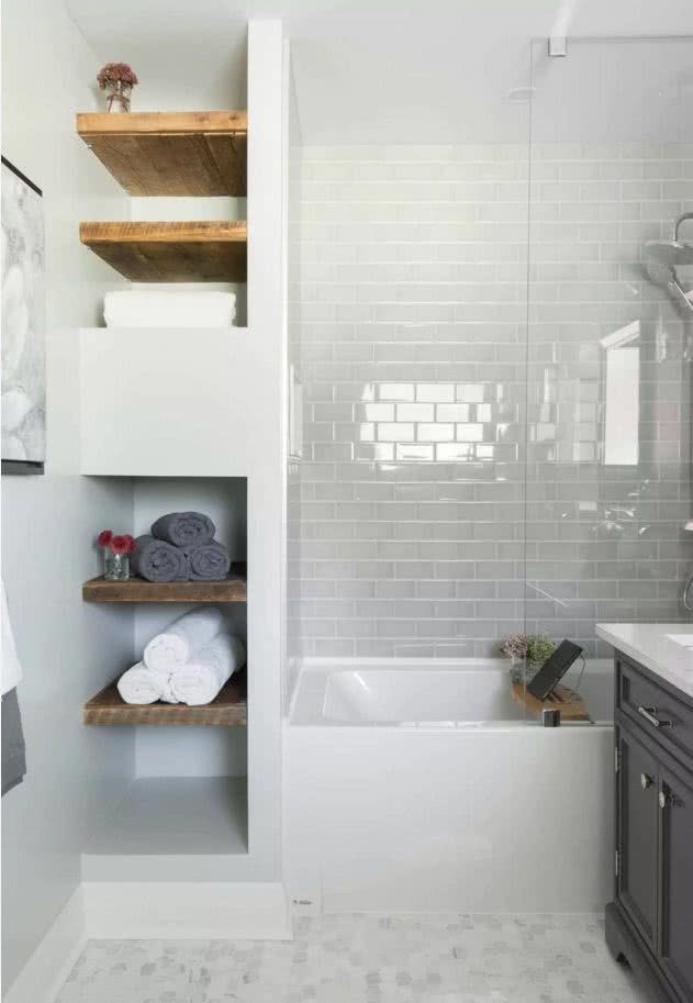 Ducha recubierta de azulejos rectangulares grises, al lado de bañera hay estantes flotantes en madera con toallas y artículos de baño