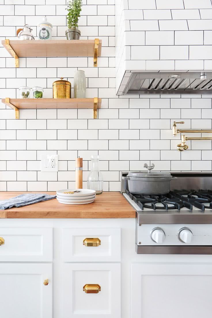 cocina con azulejos rectangulares blancos, armarios bajos blancos, encimera y estantes en madera, metales decorativos dorados