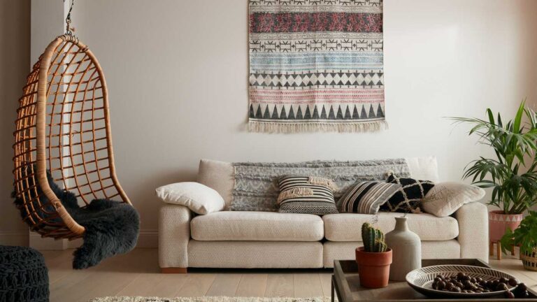 Sala con paredes blancas, sofá blanco roto con cojines étnicos y tapiz, sillón colgante de mimbre