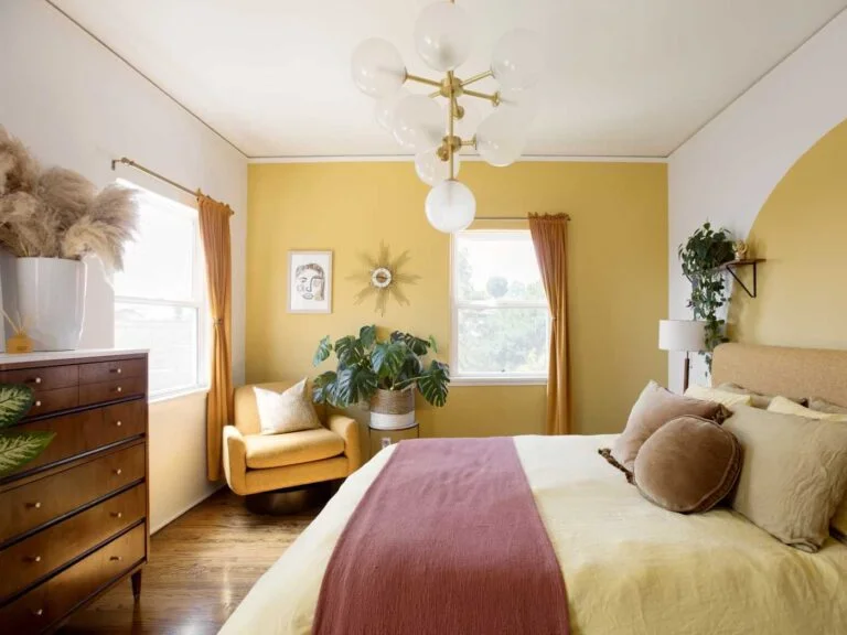 Habitación con pared amarilla, el resto blanco, suelos de madera, aparador de madera oscura, cama con cubrecamas en rojo viejo