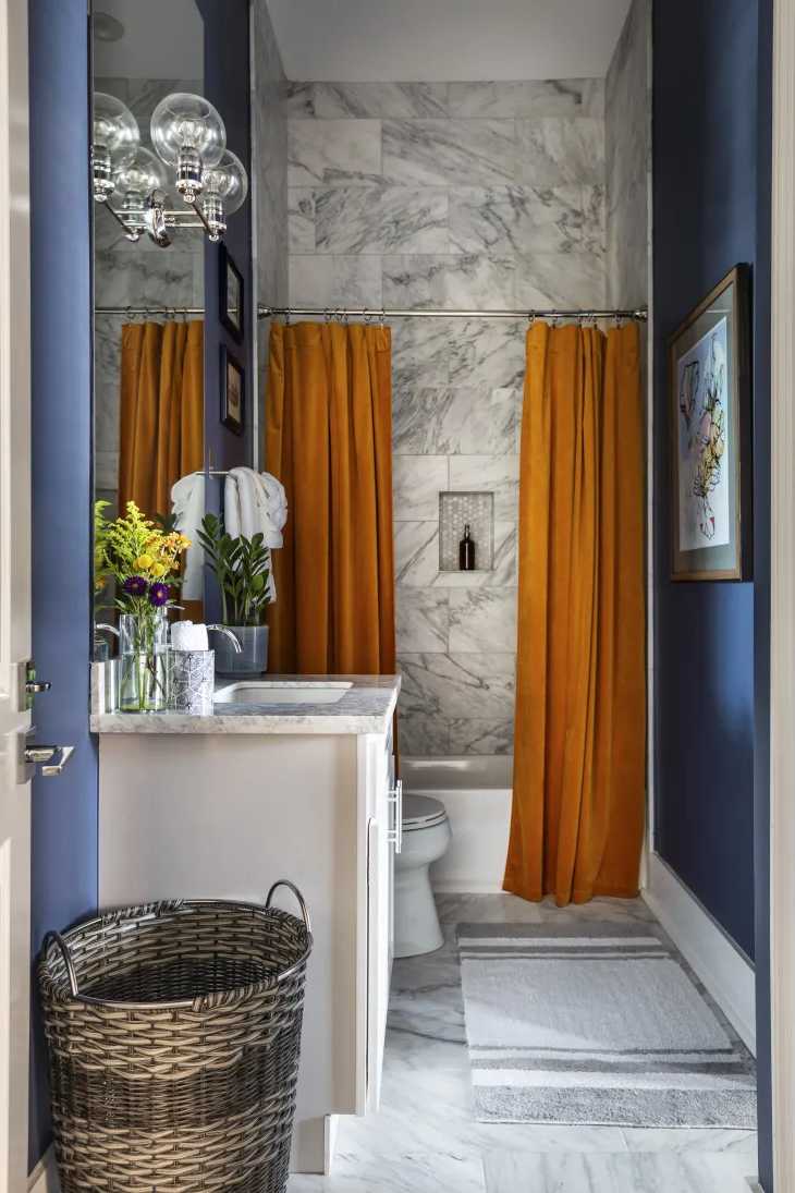Cuarto de baño con paredes azules, cortinas en tono amarillo mostaza, ducha recubierta con azulejos tipo mármol